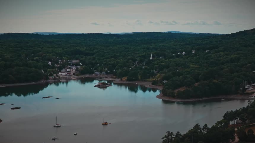 A wide aerial view slowly pulls back from a quaint coastal town nestled in a dense forest, revealing a calm bay with moored boats