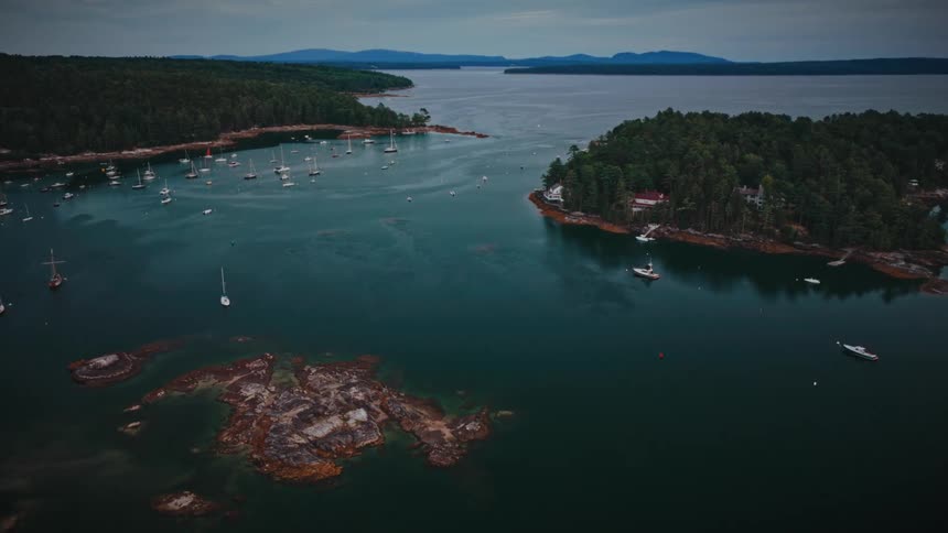 A serene aerial flyover of a tranquil harbor filled with sailboats nestled between lush, tree-covered islands on a cloudy day