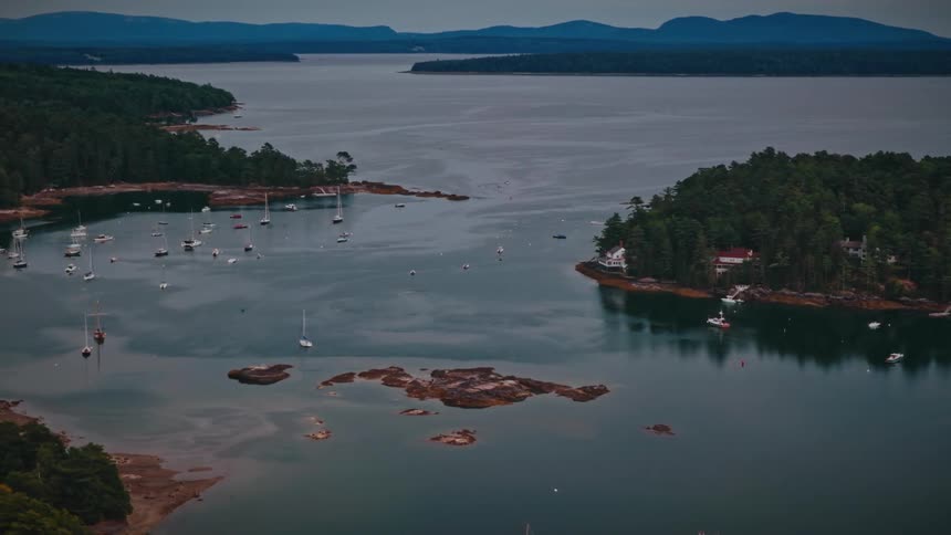 A slow aerial pullback reveals a tranquil harbor filled with sailboats nestled among rocky, forested islands on an overcast evening