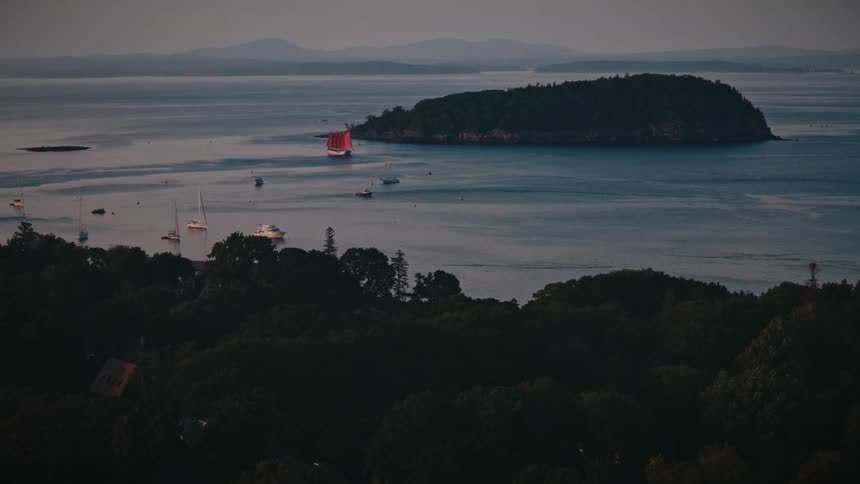 Aerial pullback from a red-sailed schooner on the open water to reveal a peaceful coastal town and harbor at dusk