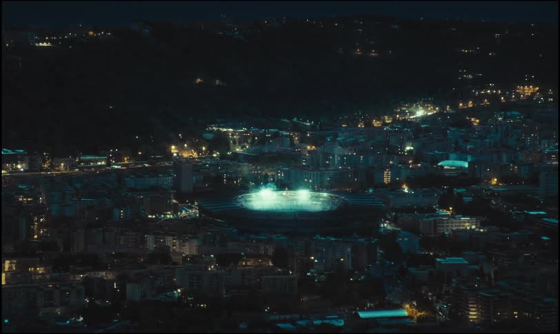 A brightly lit stadium shines in the center of a dark city nestled against a hill at night