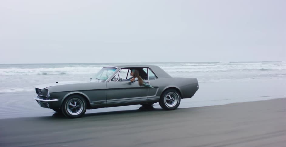 A couple takes a slow-motion drive in a classic grey Mustang along the ocean on an overcast day