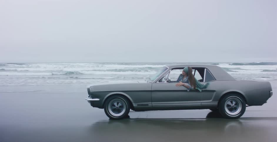 Slow motion side view of a young woman leaning out of a classic car driving along the shoreline on a moody, overcast day