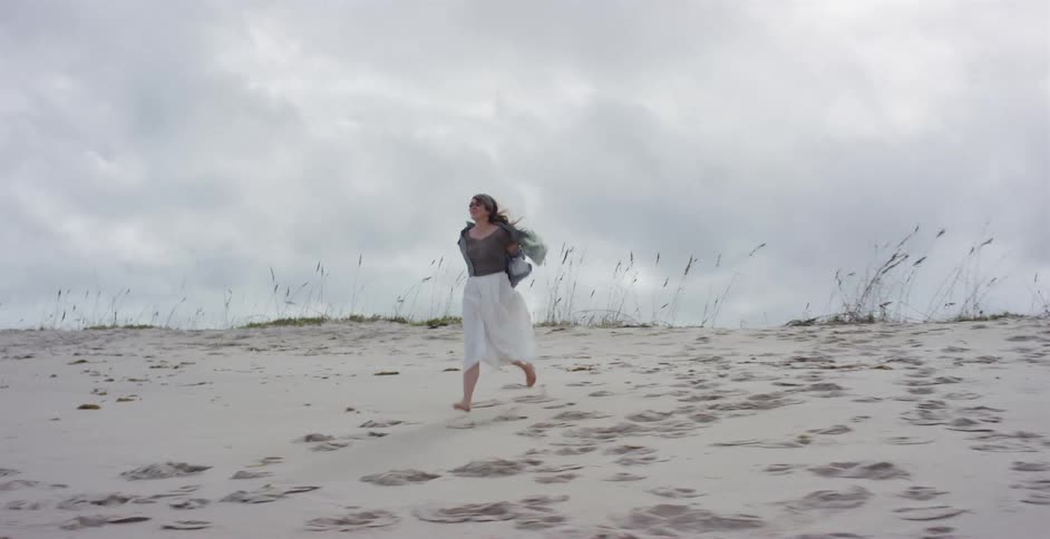 A joyful woman with long hair runs barefoot in slow motion towards the camera across a sandy beach dune on a windy, overcast day
