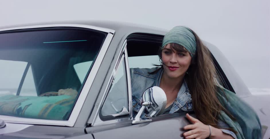 A beautiful woman smiles as she feels the breeze from the passenger window of a classic car driving along the beach