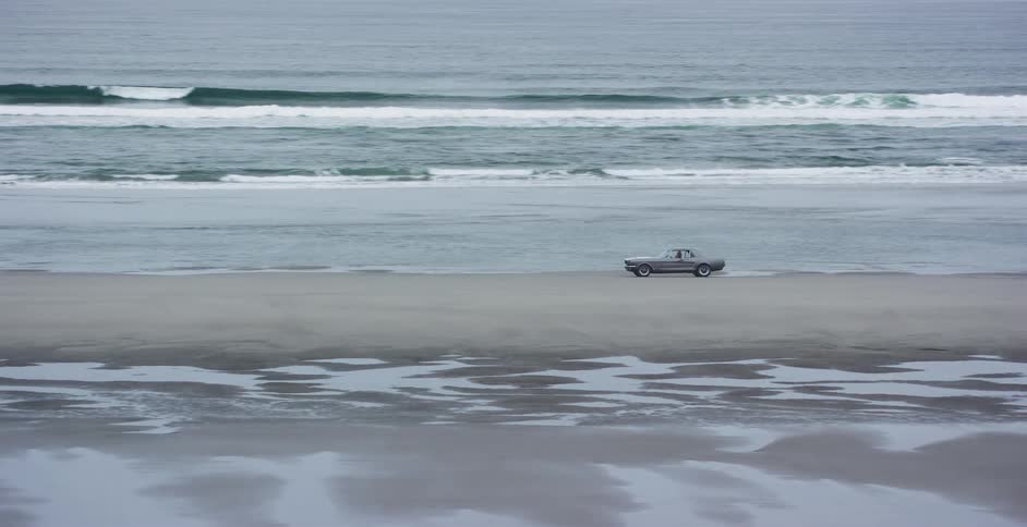 A vintage car drives along the shoreline of a vast, empty beach on an overcast day