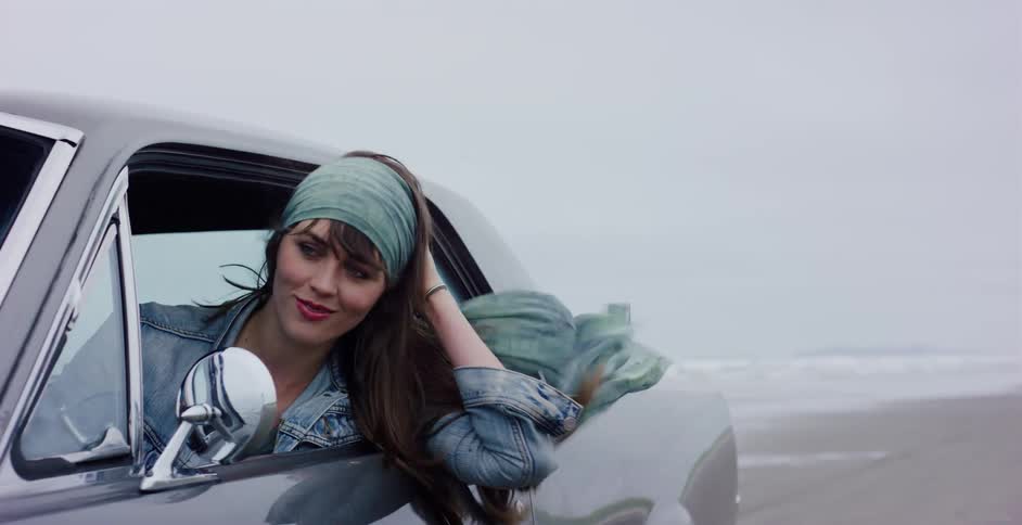 A stylish young woman leans out of a classic car, feeling the wind in her hair and scarf as she enjoys a scenic drive along the ocean beach