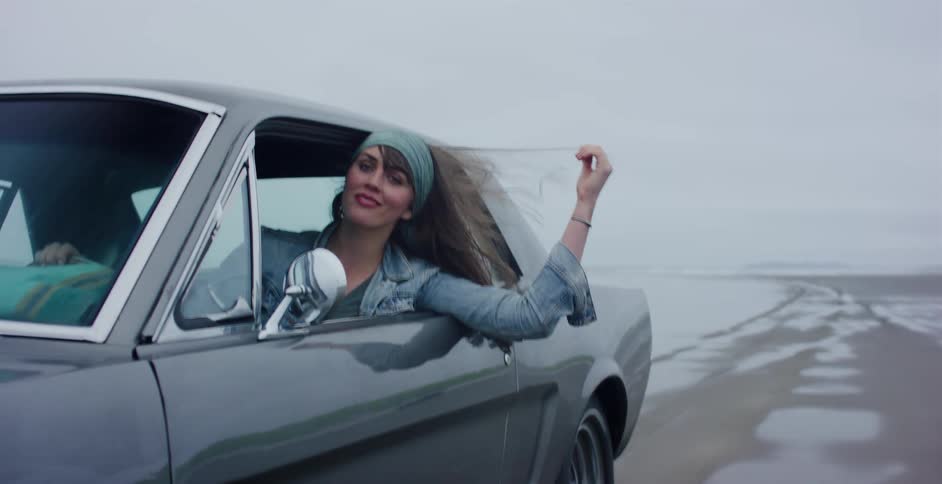 A free-spirited woman leans out of a vintage muscle car, smiling and playing with her hair in slow motion as it blows in the wind on a deserted beach