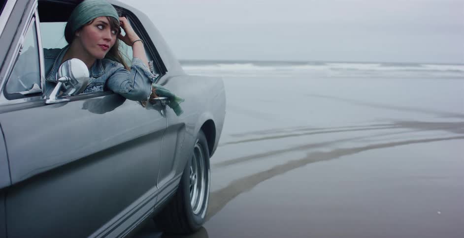 A thoughtful woman leans out of her classic car window to watch the ocean on a cloudy day at the beach