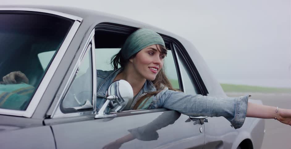 A carefree young woman smiles from the driver's seat of a classic car with her arm out the window enjoying the ride