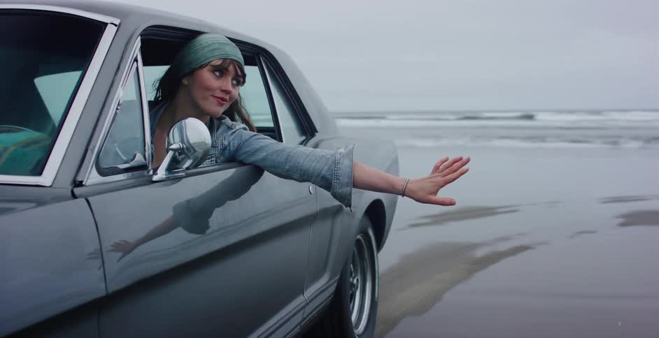 A carefree woman in a classic car rides along the ocean beach with her arm out the window, enjoying the ride