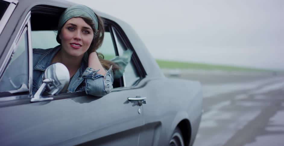 A young woman in a denim jacket leans out the window of a classic muscle car as it drives down a wet road on an overcast day