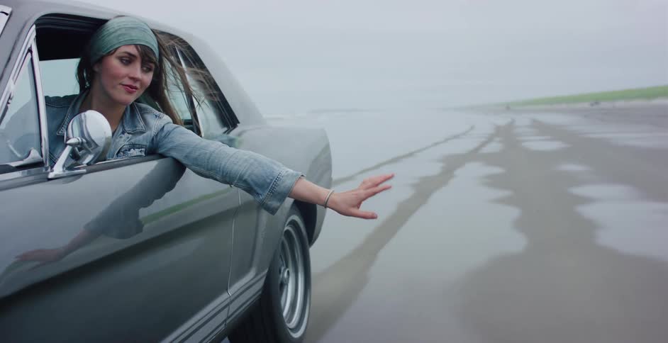 A free-spirited woman feels the wind on her hand while cruising in a classic car along a misty, wet beach