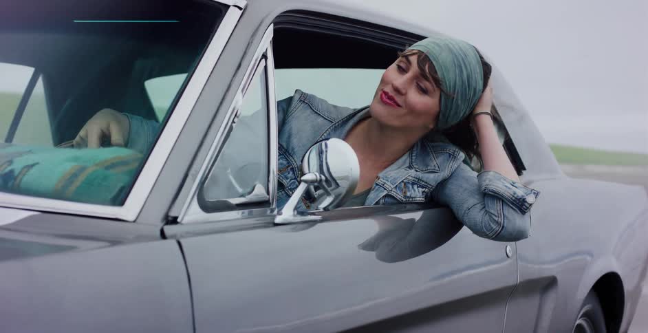 A carefree woman enjoys the wind blowing through her hair as she leans out the window of a classic grey Mustang on a road trip
