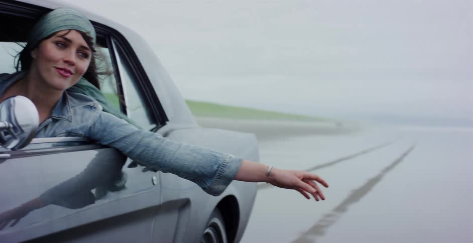 A free-spirited woman enjoys the ocean breeze with her arm out the window while driving in a classic car along a foggy beach