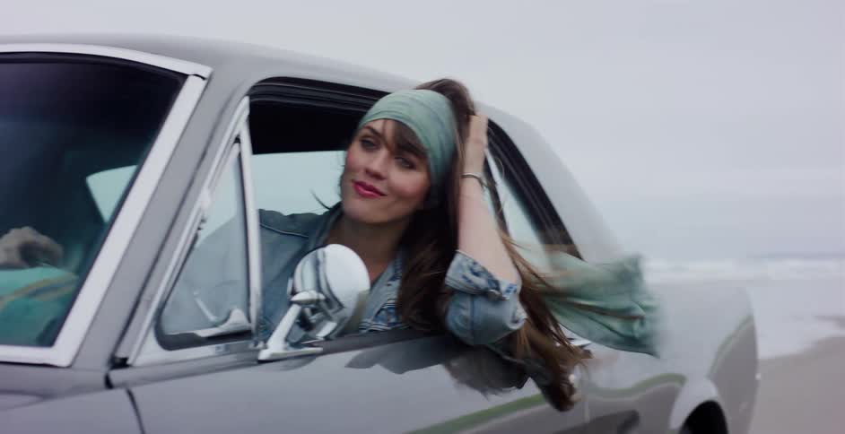 A beautiful young woman leans out of a classic Mustang's window, enjoying the breeze as the car drives along a foggy beach