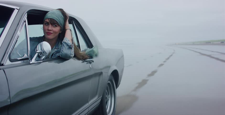 A cool and confident woman with a headscarf looks out the window of a classic muscle car while driving on a foggy, wet beach