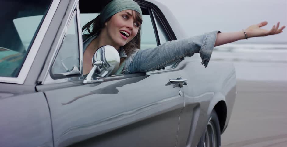 A carefree woman leans out of a classic car window, feeling the wind in her hand as she drives along a sandy beach