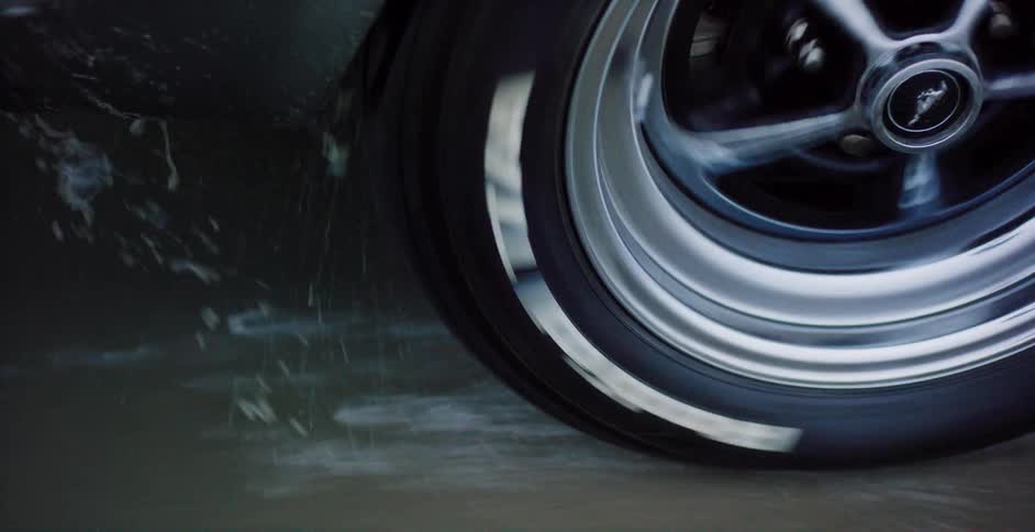 The spinning whitewall tire of a classic Ford Mustang kicks up water as it performs a burnout on wet asphalt