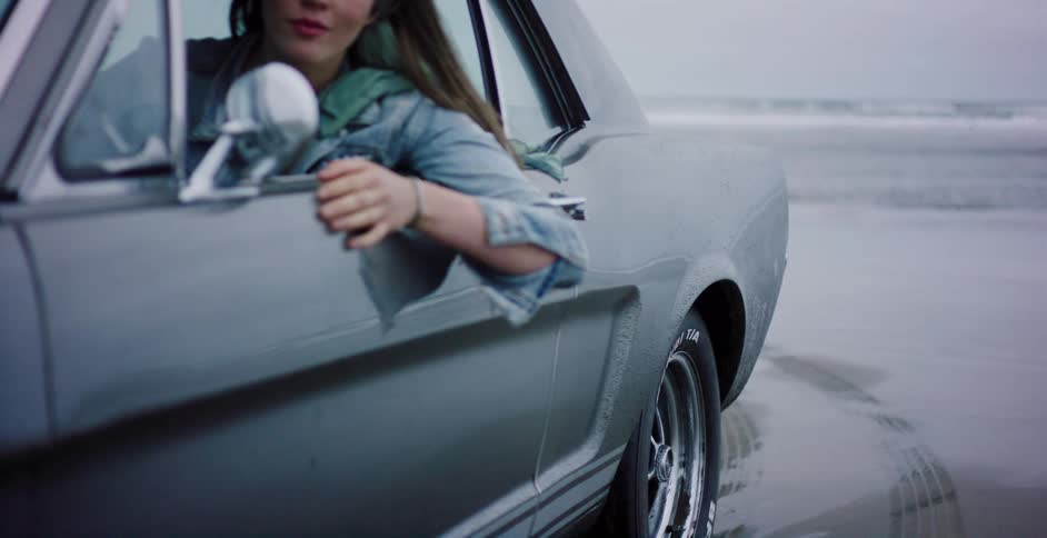 Close up on a classic grey car parked on a wet beach with a young woman in the driver's seat looking out at the ocean
