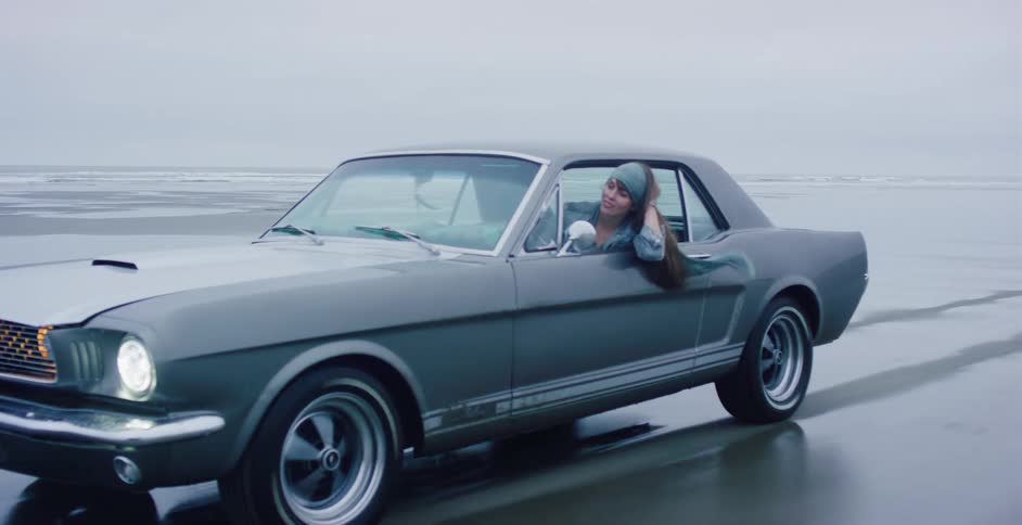 A stylish woman with long hair flowing in the wind drives a classic grey Mustang along the wet sand of an ocean beach