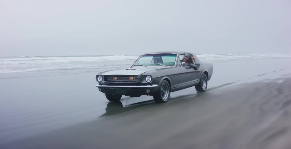 A carefree woman leans out of a vintage Ford Mustang as it cruises along a wet, sandy beach on a moody, overcast day