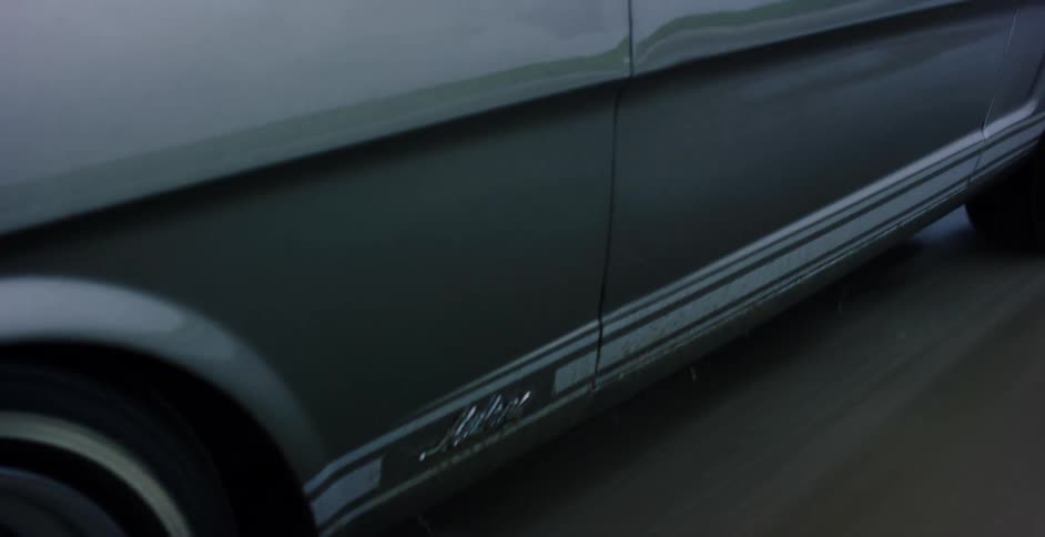 A close-up shot of a classic muscle car's spinning wheel pulls back to reveal a stylish woman driving on a misty beach
