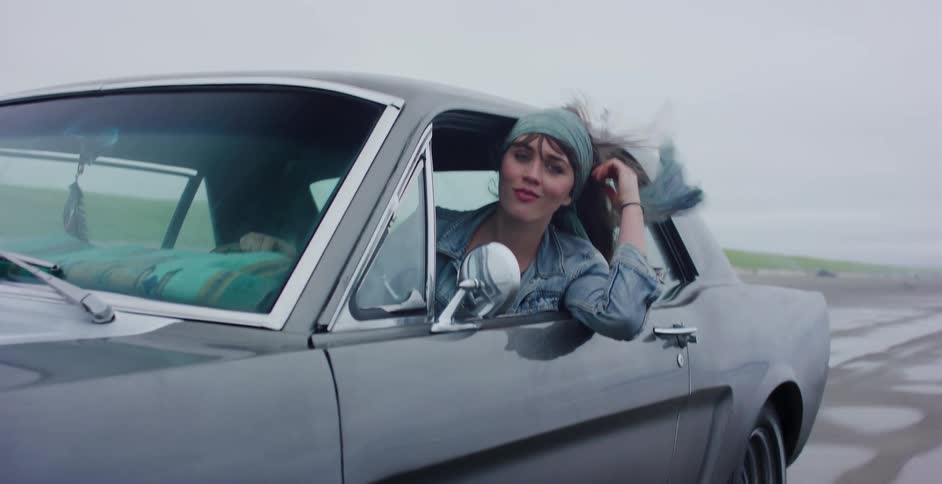 A stylish young woman leans out the window of her classic grey Ford Mustang in slow motion, her hair blowing in the wind as she cruises down an open road