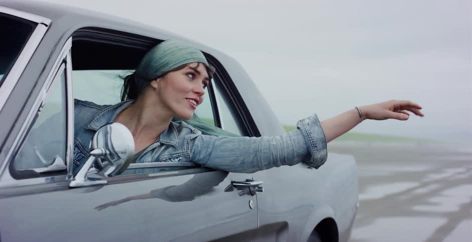 A free-spirited woman in a classic car smiles as she holds her arm out the window, her headscarf blowing in the wind on a foggy day