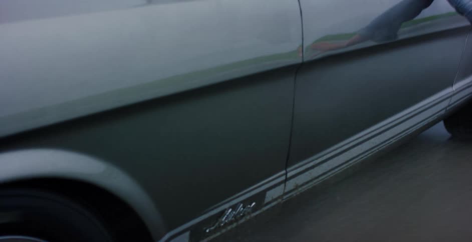 A close-up shot of a classic Ford Mustang's wheel transitions to a reveal of a young woman enjoying a drive on a wet beach