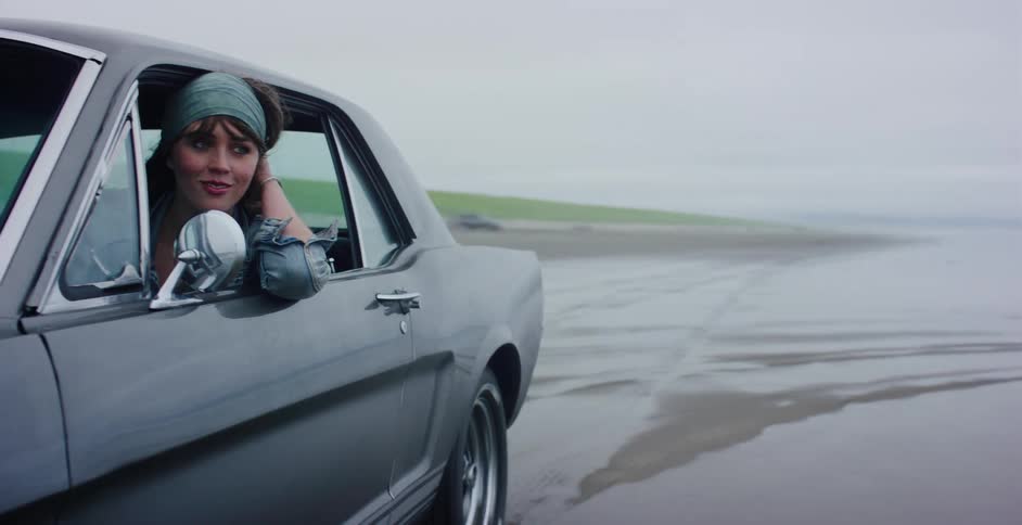 A young woman leans out the window of her classic muscle car as she drives along a wet sandy beach on an overcast day
