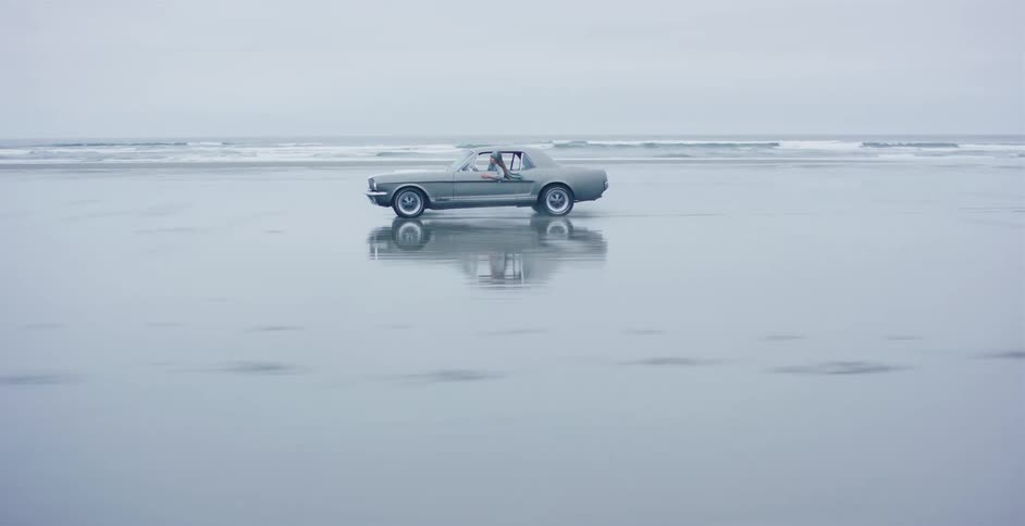 A classic Ford Mustang drives along the reflective wet sand of the beach on a moody, overcast day