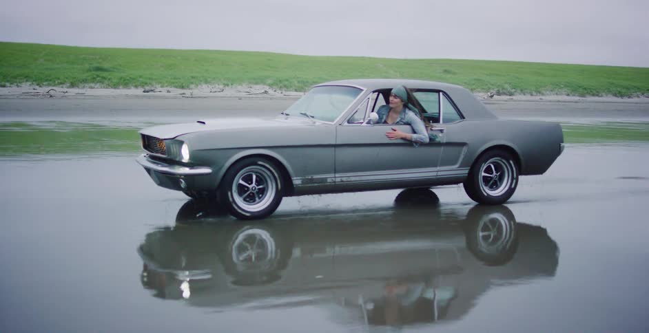 Cinemagraph of a woman in a classic grey Mustang stopped in the shallow ocean water on a beach, with its wheels spinning and splashing