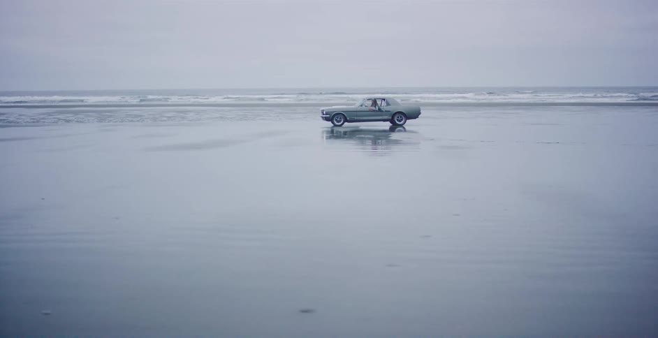 A classic green muscle car is parked on a vast, wet beach, reflecting perfectly on the sand under an overcast sky