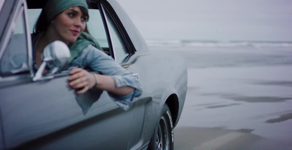 A stylish young woman enjoys a slow-motion drive in a vintage car along the wet, sandy shore of the ocean on an overcast day