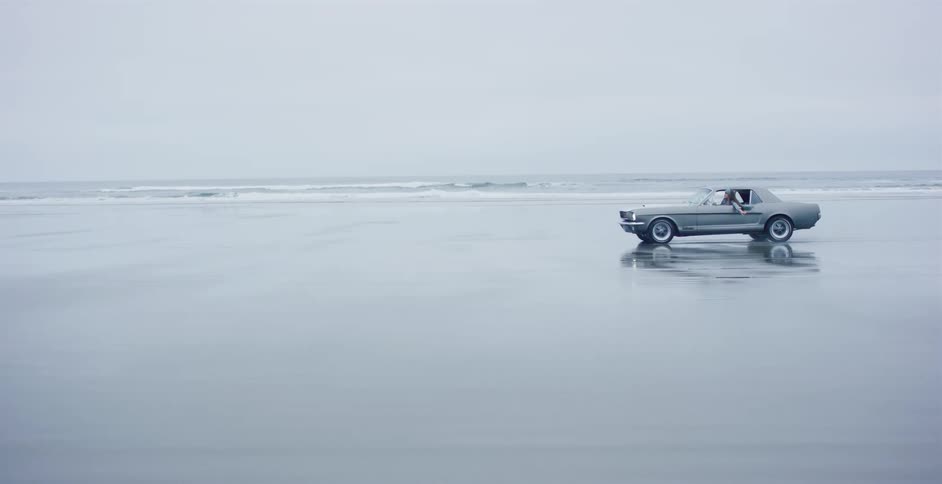 A tracking shot follows a classic silver car as it cruises along a vast, wet beach next to the ocean on an overcast day