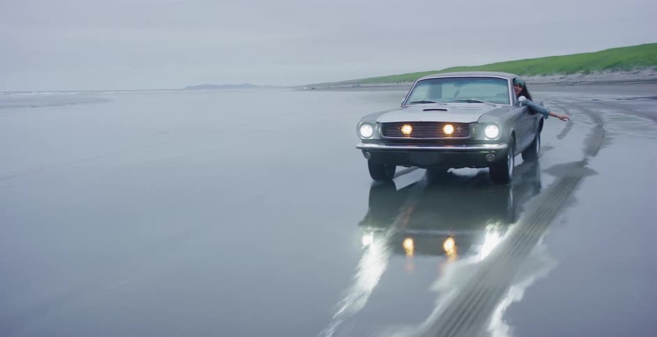 A woman leans out the window of a classic silver Ford Mustang as it drives on the wet sand of a remote beach on an overcast day