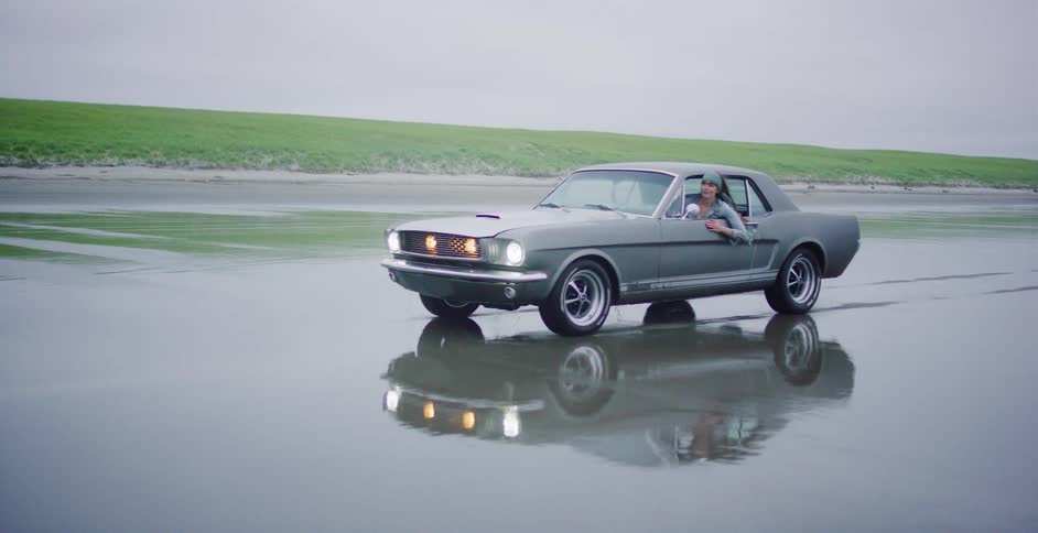 A classic grey Ford Mustang drives slowly through the shallow water of a beach with a grassy dune in the background