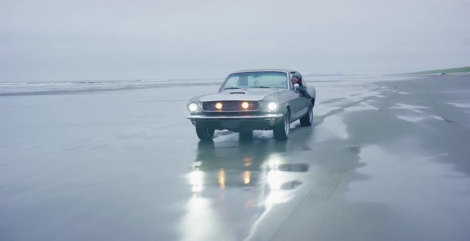 A woman hangs out the window of a classic Ford Mustang as it drives along the wet, reflective sand of an empty beach on a foggy day