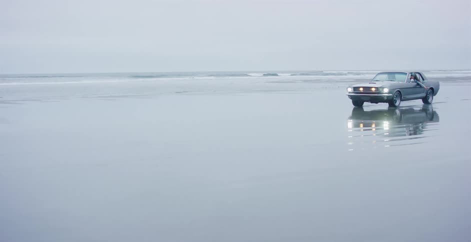 A classic muscle car slowly drives along the edge of the ocean on a vast, reflective beach on an overcast day