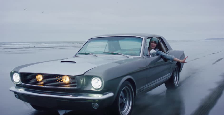 A stylish young woman hangs out the passenger side window of a classic muscle car as it drives along a wet beach in slow motion