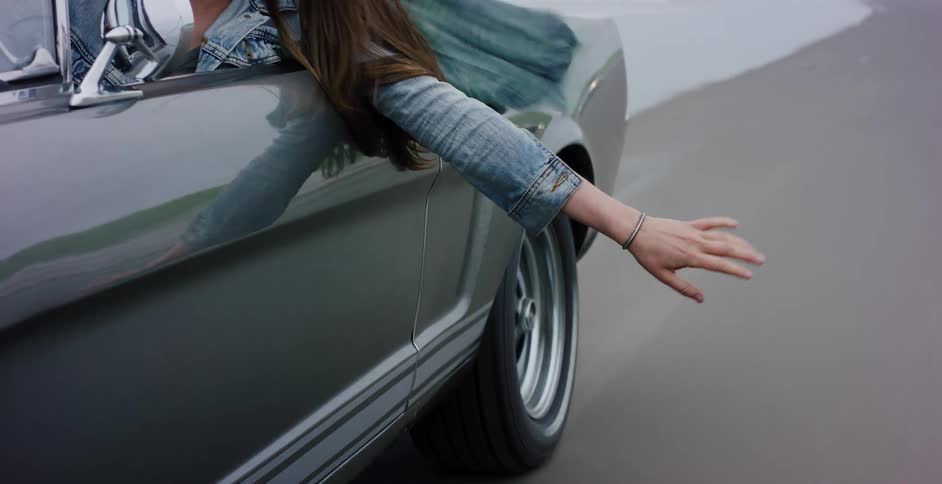 Slow motion close-up of a woman's hand feeling the wind as she drives a classic car along a wet sandy beach
