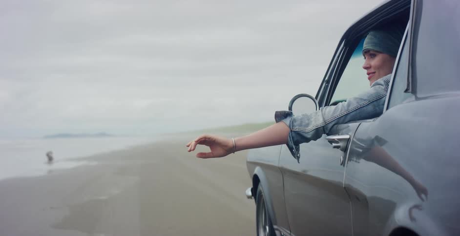 A woman's arm reaches out from the passenger window of a vintage car as it drives on a sandy beach, scattering seagulls in slow motion