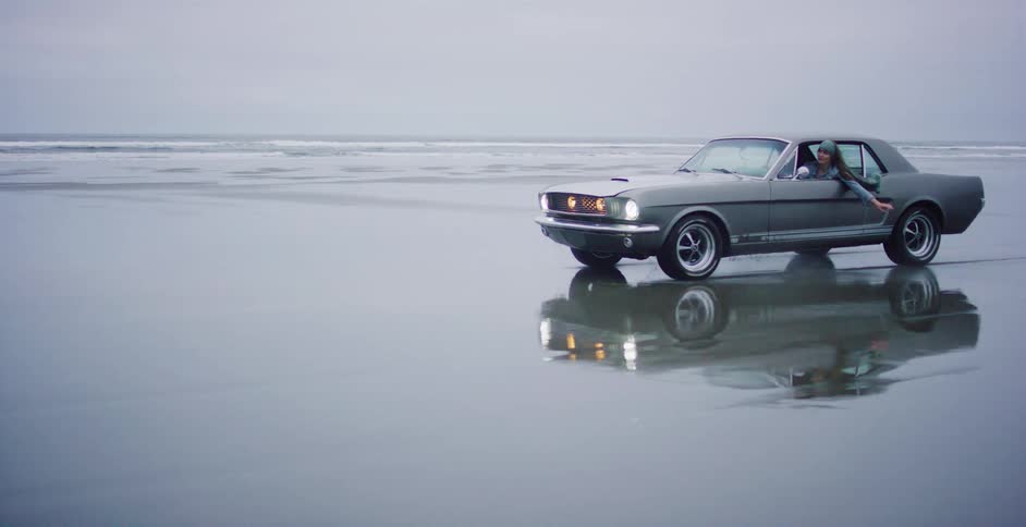 A woman in a classic grey Mustang drives slowly along a wet, reflective beach, leaning out the window with her hair blowing in the wind