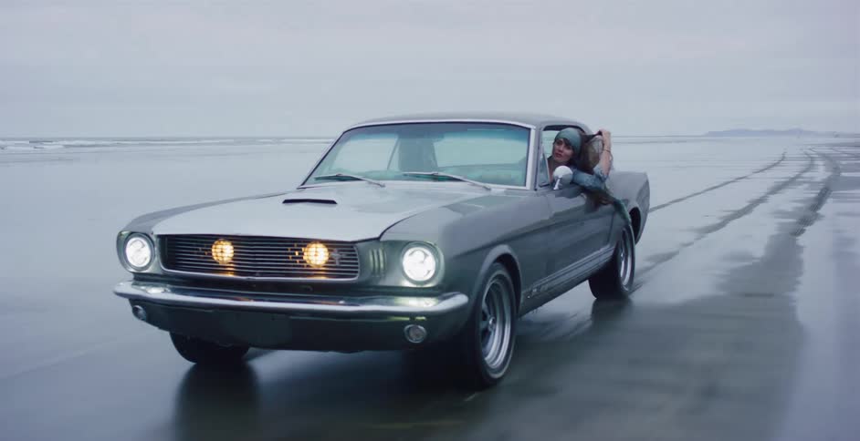 A young woman hangs out the window of a classic Ford Mustang as it drives along a wet beach on a cool, overcast day