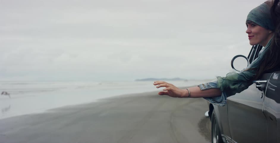 A free-spirited woman holds her hand out the window of a moving car as seagulls fly alongside on an overcast beach in slow motion