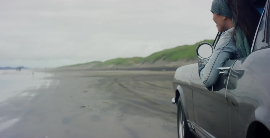 A beautiful young woman with her hair blowing in the wind enjoys a scenic drive in a classic car along a sandy beach in slow motion