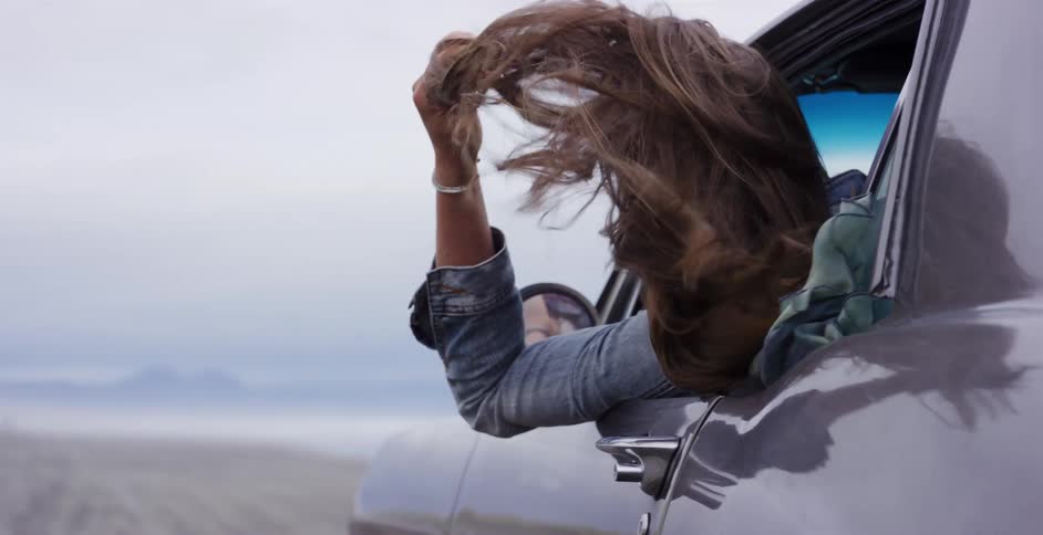 A free-spirited woman hangs out of a classic car window, her long brown hair blowing in the wind in slow motion