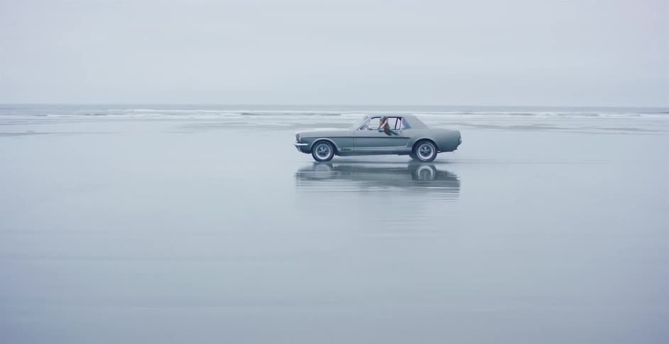 A vintage grey Mustang cruises across a vast, wet beach, its reflection shimmering on the sand under an overcast sky