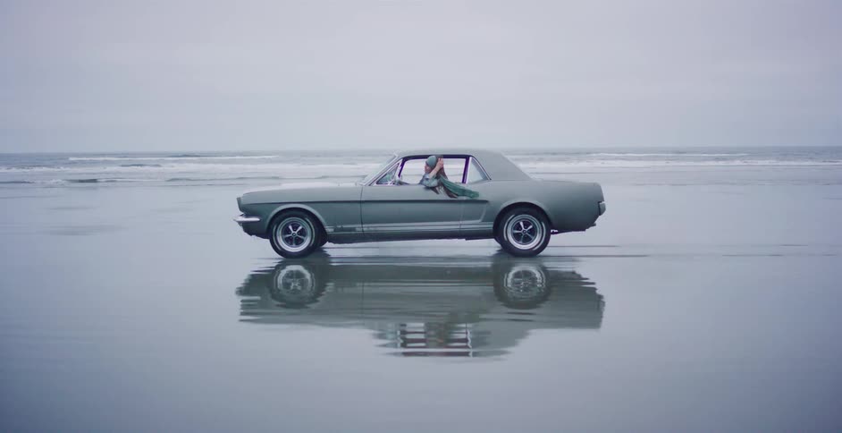 A classic Ford Mustang is parked on a wet, sandy beach, its reflection perfectly mirrored in the water as a person leans out the window